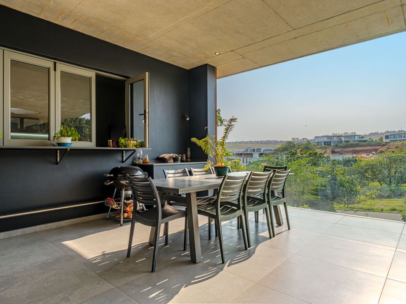 A modern covered patio featuring tiled flooring, a concrete ceiling, and a glass balustrade with expansive views, including a convenient pass-through window to the kitchen.