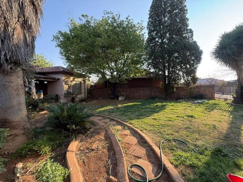 A spacious backyard garden featuring mature trees, a brick boundary wall, and a paved pathway leading towards the rear of the house.