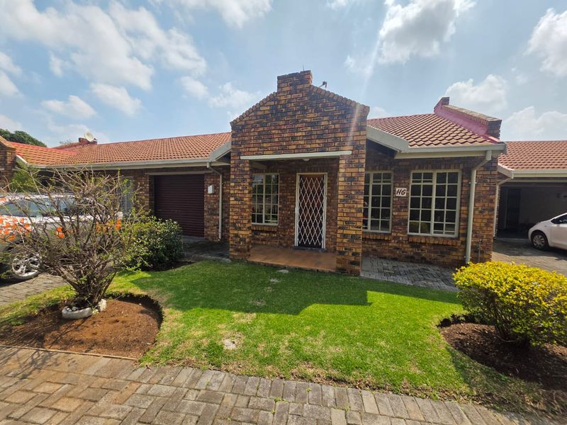 Front elevation of a face brick townhouse featuring a tiled roof, security entrance gate, paved driveway, and a well-maintained front garden.