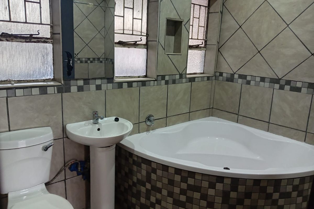 A dated bathroom featuring a corner bathtub, pedestal basin, and toilet with tiled finishes and security bars on the windows.