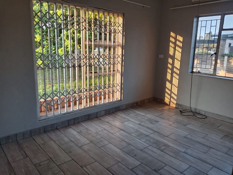 An empty living area featuring modern wood-look floor tiles, security gates on windows, and an air conditioning unit, bathed in natural afternoon light.