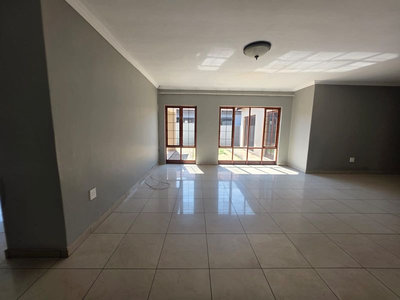 A spacious, empty living area featuring neutral grey walls, large tiled flooring, and double glass doors providing natural light and access to the exterior.