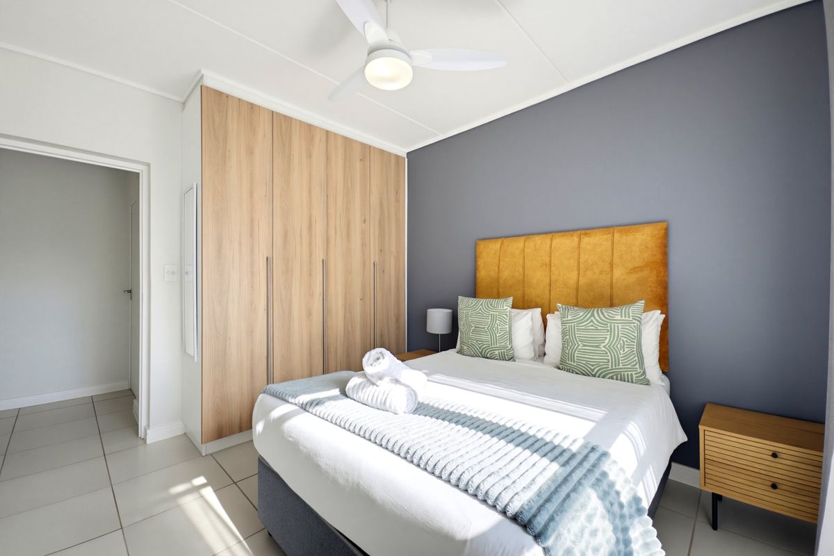 A well-maintained bedroom featuring light wood-finish built-in cupboards, a modern ceiling fan, and neutral floor tiles with a contrasting feature wall.
