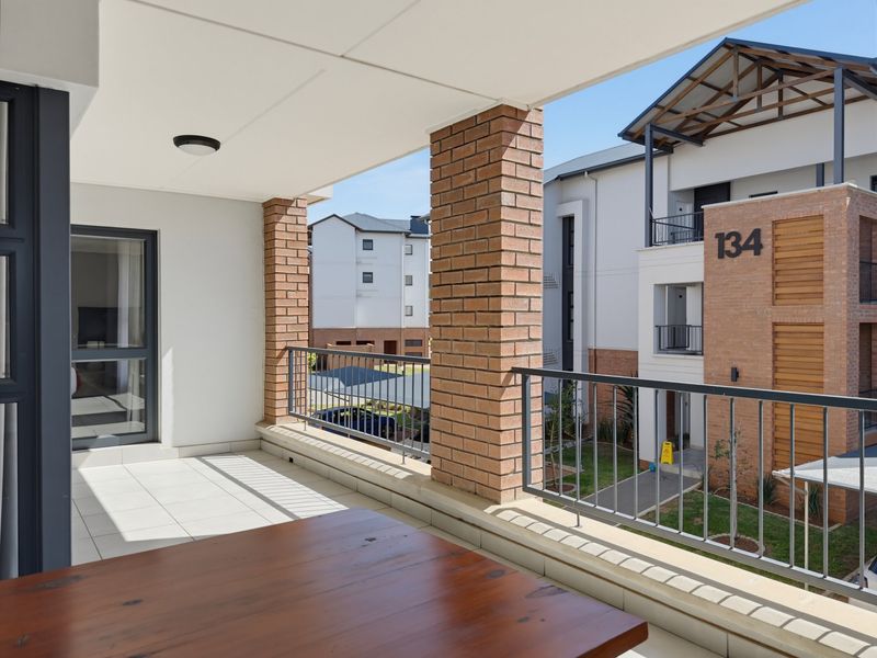 A spacious covered balcony featuring tiled flooring, brick support pillars, and a metal balustrade, overlooking the apartment complex grounds.