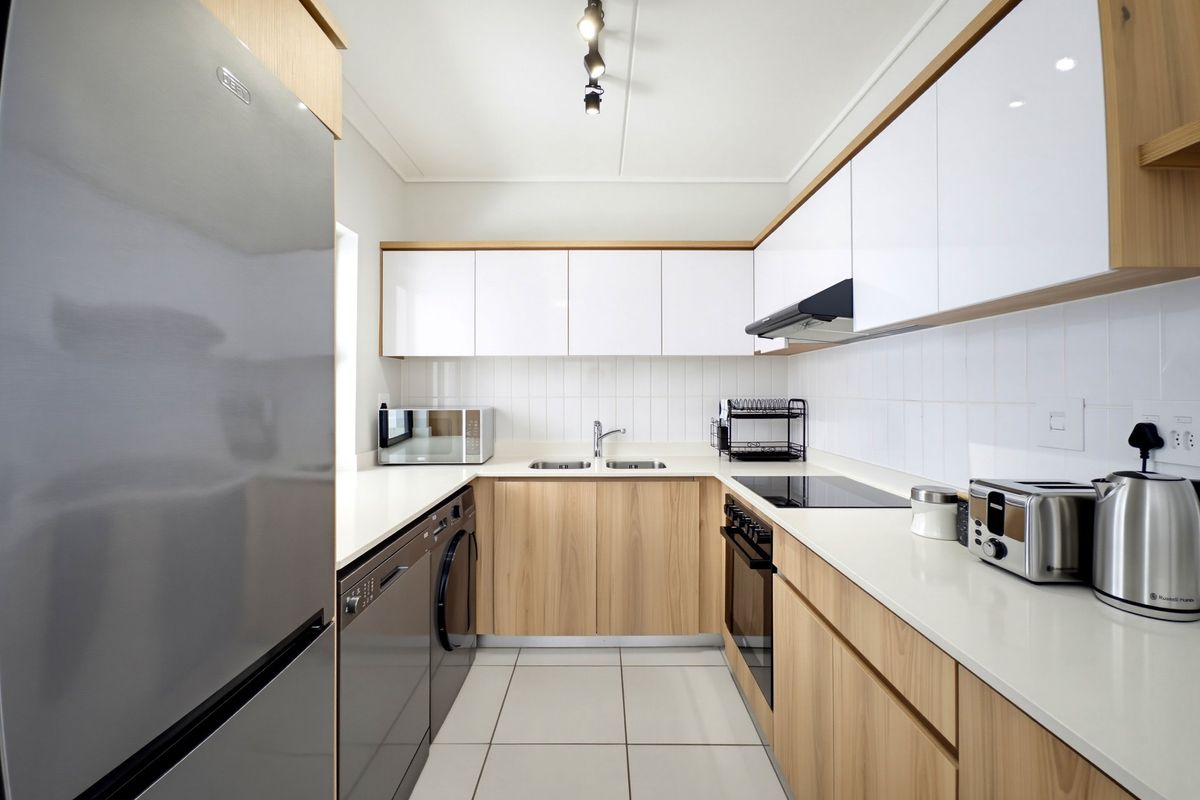 Modern galley-style kitchen featuring light wood-grain cabinetry, white high-gloss upper cabinets, quartz countertops, and integrated appliances with a clean subway tile backsplash.