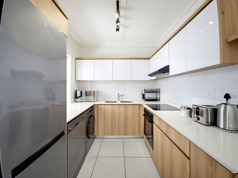Modern galley-style kitchen featuring light wood-grain cabinetry, white high-gloss upper cabinets, quartz countertops, and integrated appliances with a clean subway tile backsplash.