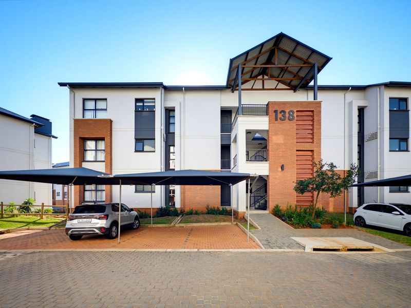 Modern apartment building exterior featuring brick and plaster facade, covered parking bays, and a paved entrance area.