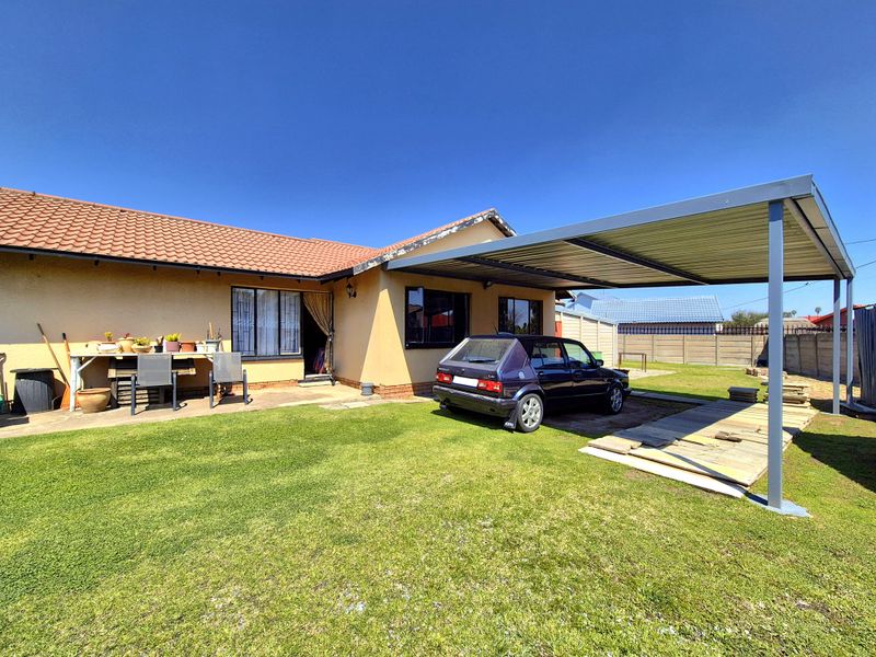 Rear exterior view of the house featuring a paved patio area, a metal carport structure, and a spacious garden with perimeter walling.