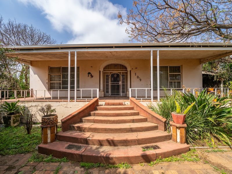 Front elevation of a single-story house featuring a covered veranda, concrete entrance stairs, and established garden surroundings.