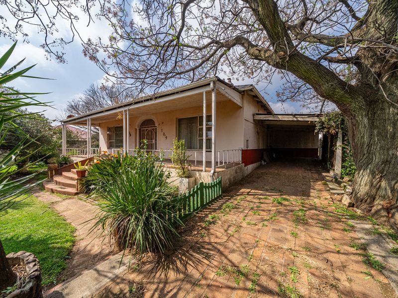 Front elevation of a single-story house featuring a covered porch, brick driveway, and attached carport, set within a mature garden.