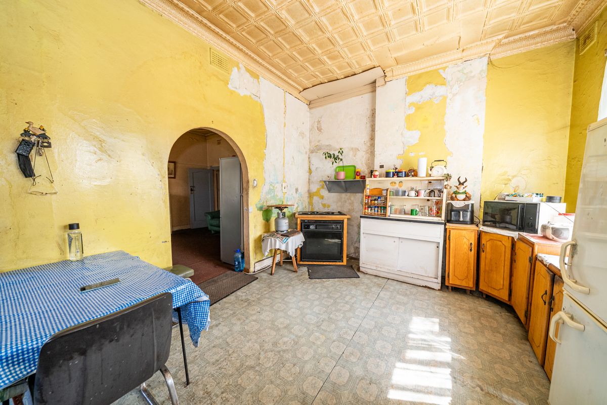 A dated kitchen featuring a pressed metal ceiling, vinyl flooring, and an archway, currently requiring significant renovation and cosmetic repairs to the walls and ceiling.