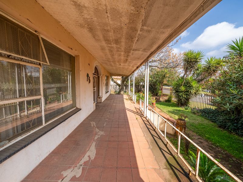 A covered outdoor patio featuring tiled flooring and metal railings, overlooking a garden area. The space shows signs of wear and requires maintenance.