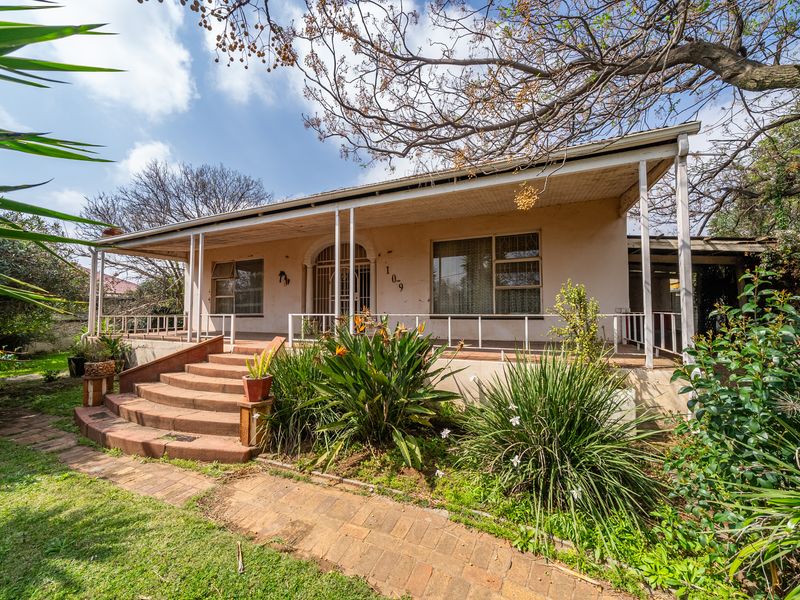 Front elevation of a single-story house featuring a covered veranda, brick staircase, and established garden surroundings.