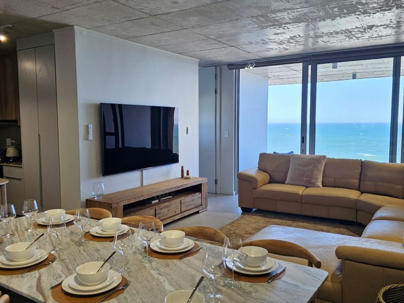 Modern open-plan living and dining area featuring an exposed concrete ceiling, large sliding glass doors, and expansive ocean views.
