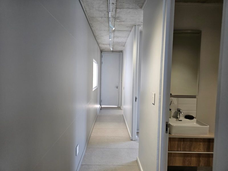 A modern hallway featuring an exposed concrete ceiling with track lighting, tiled flooring, and access to a bathroom with a contemporary vanity.