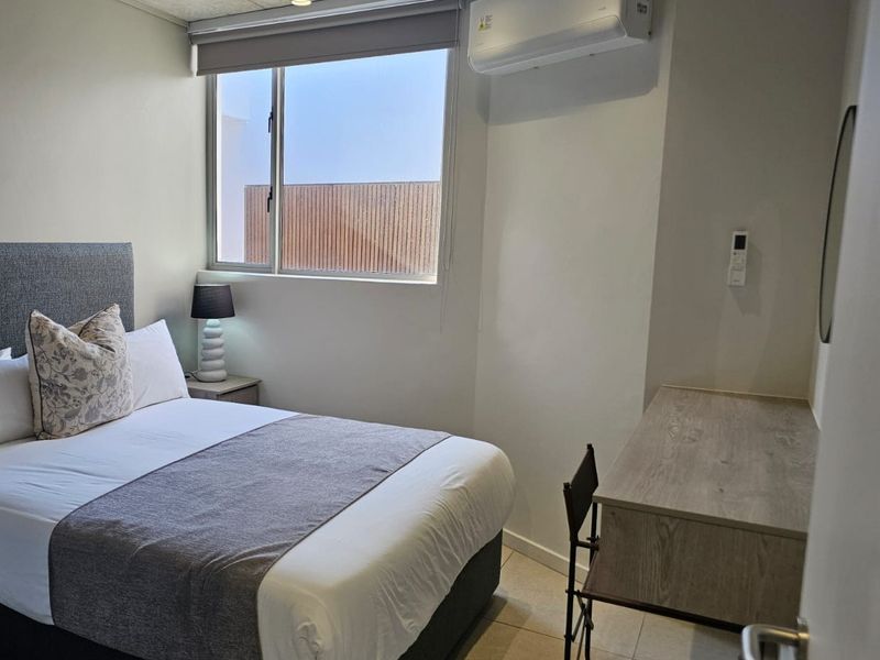 A modern bedroom featuring an exposed concrete ceiling, tiled flooring, air conditioning, and a large window with a roller blind.