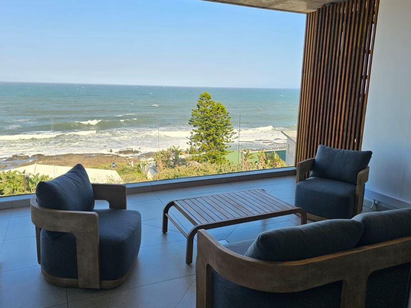 A modern covered balcony featuring a glass balustrade, exposed concrete ceiling, and timber slat detailing, offering expansive ocean views.