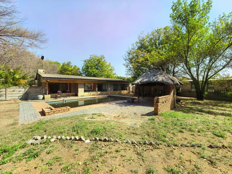 Rear exterior view featuring a swimming pool, brick paved patio area, and a traditional thatch lapa set within a large garden.