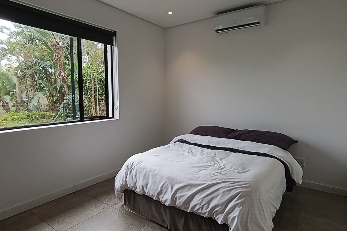 A modern bedroom featuring neutral tiled flooring, recessed downlighting, and a wall-mounted split-system air conditioner with views of the garden.
