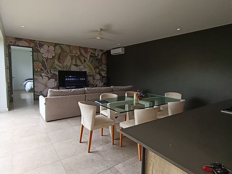 Open-plan living and dining area featuring tiled floors, a kitchen island, ceiling fan, and split-system air conditioning with a decorative feature wall.