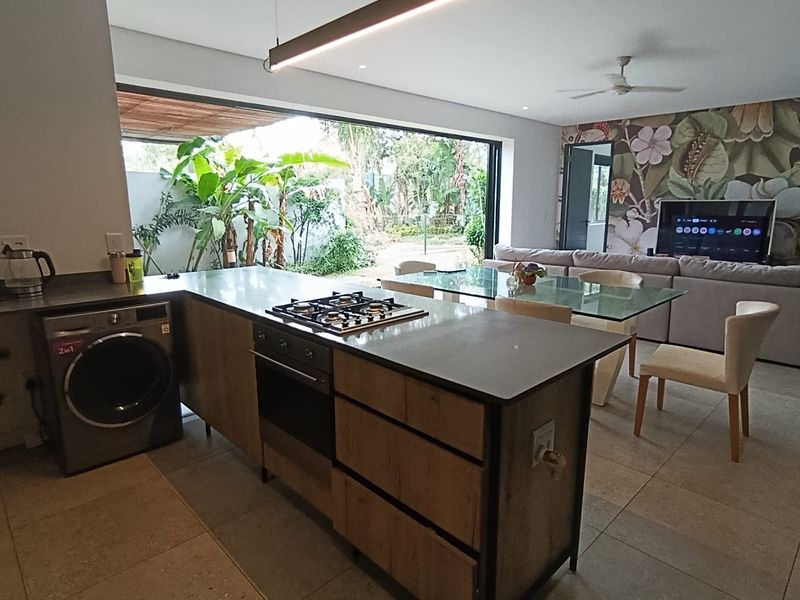 Modern open-plan living area featuring a central kitchen island with built-in hob and oven, flowing seamlessly to an outdoor patio through large sliding doors.