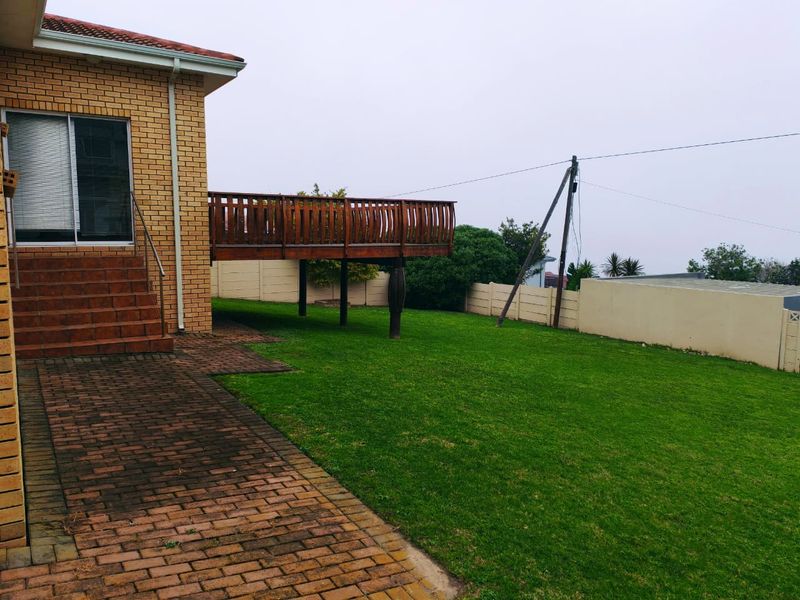 Rear exterior view featuring a raised wooden deck, brick paved patio area, and a spacious lawn enclosed by a boundary wall.