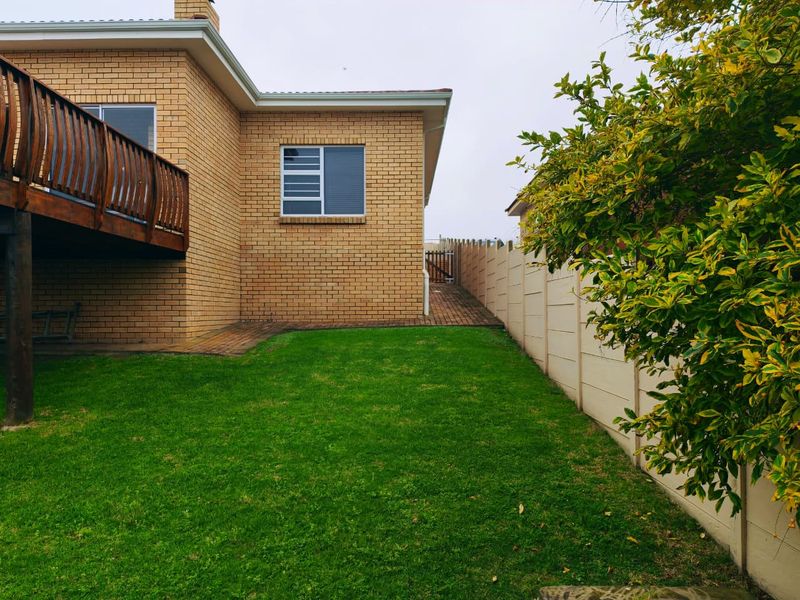 Rear view of the property featuring a brick facade, a raised wooden deck, a well-maintained lawn, and a secure precast concrete boundary wall.