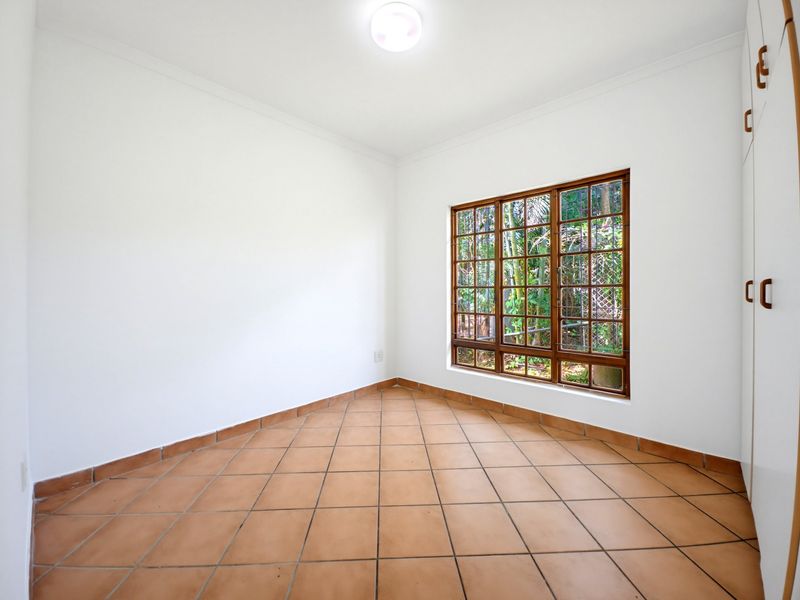 An empty bedroom featuring terracotta floor tiles, built-in cupboards, and a large wooden window with security bars overlooking the garden.