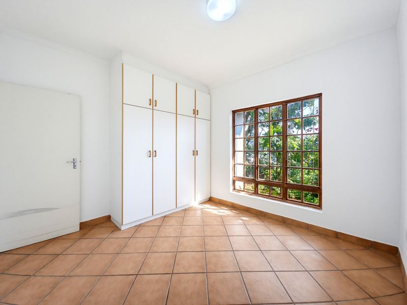A bright, empty bedroom featuring tiled flooring, built-in white cupboards, and a large wooden-framed window with security bars.