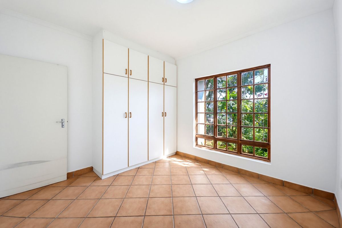 A bright, empty bedroom featuring tiled flooring, built-in white cupboards, and a large wooden-framed window with security bars.
