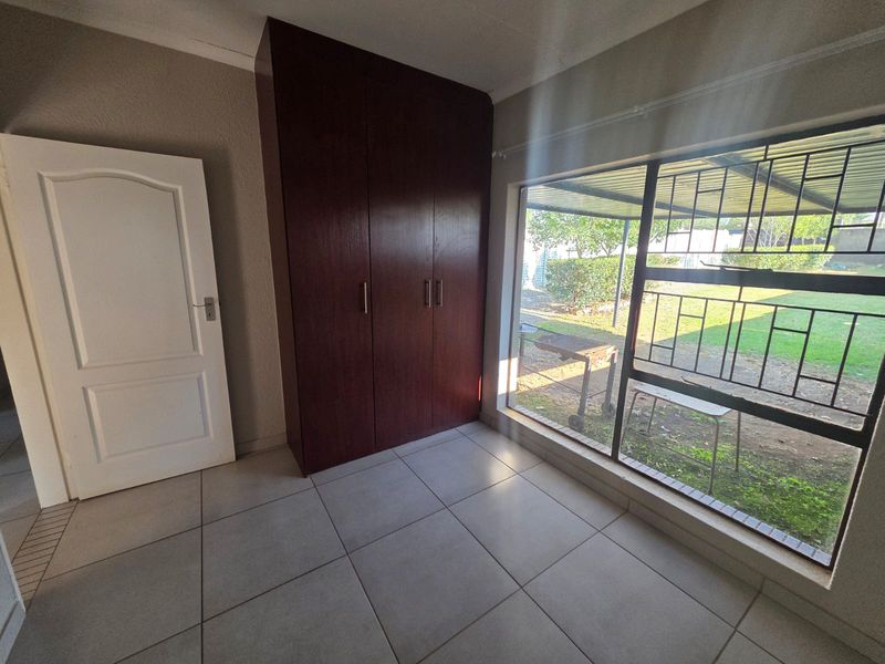A bright, empty bedroom featuring tiled flooring, a built-in wooden wardrobe, and a large window with security bars overlooking the garden.