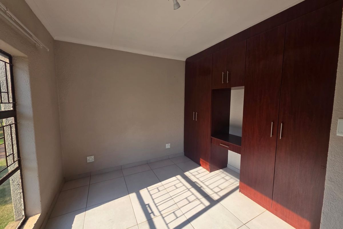 A bright, empty bedroom featuring neutral tiled flooring and dark wood built-in cupboards with an integrated desk space.