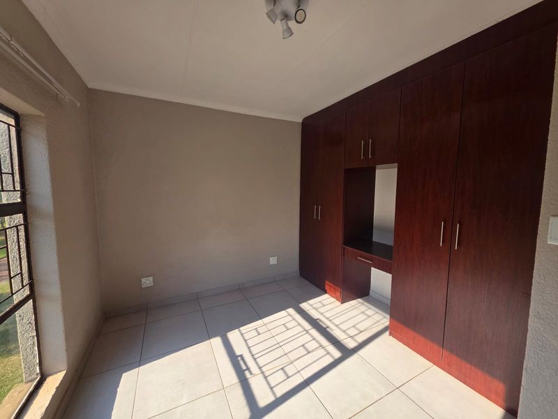 A bright, empty bedroom featuring neutral tiled flooring and dark wood built-in cupboards with an integrated desk space.