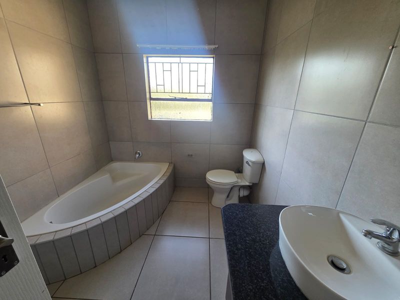 A clean, tiled bathroom featuring a built-in corner bathtub, a modern vanity basin with a granite countertop, and a toilet.