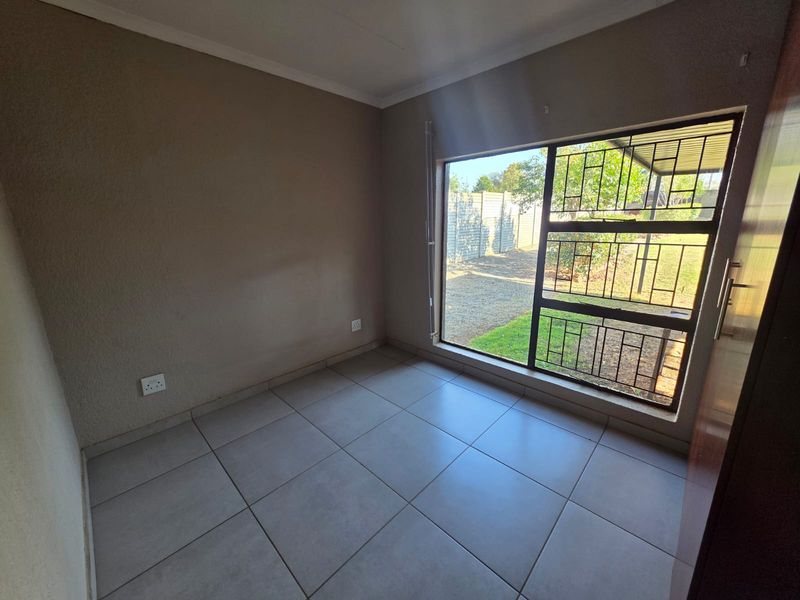 A bright, empty bedroom featuring neutral tiled flooring and a large window with security bars, offering a view of the rear garden area.