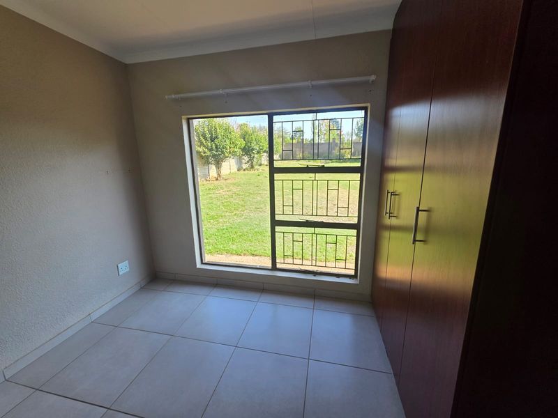 A bright, empty bedroom featuring tiled flooring, built-in wooden cupboards, and a large window with security bars overlooking the garden.
