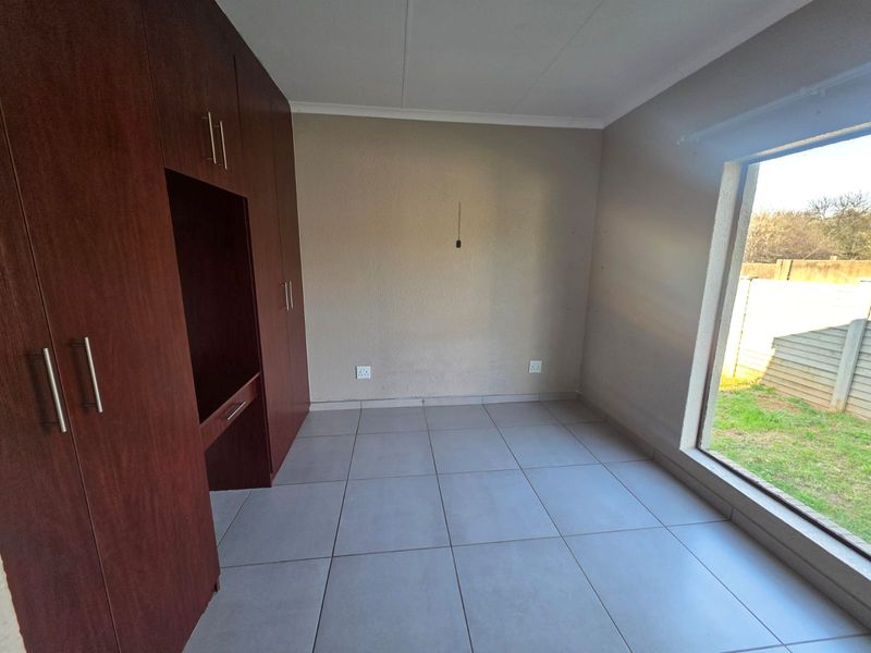 A bright, empty bedroom featuring built-in wooden cupboards, neutral tiled flooring, and a large window overlooking the garden area.