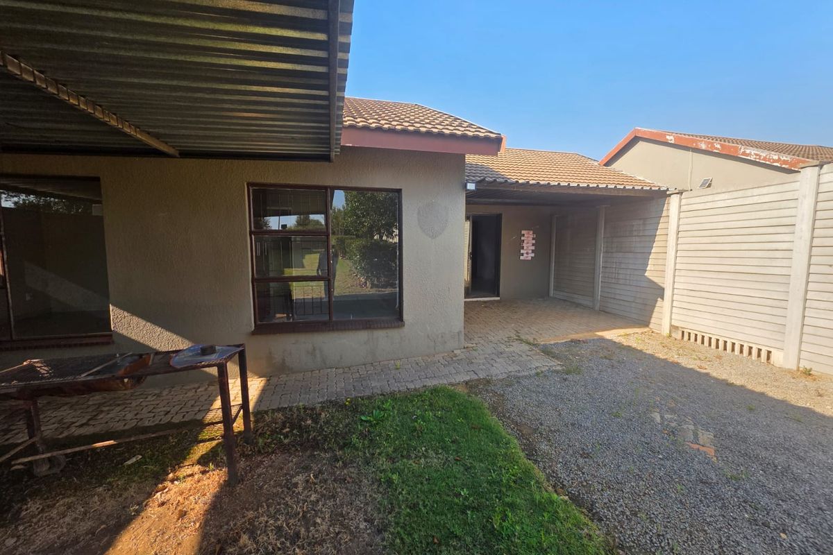 Rear exterior view of the townhouse featuring a paved patio area, corrugated metal roofing, and a gravel driveway enclosed by a precast concrete boundary wall.