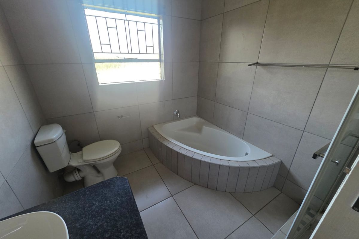 A functional bathroom featuring a built-in corner bathtub, toilet, and vanity with a dark countertop, finished with neutral wall and floor tiles.