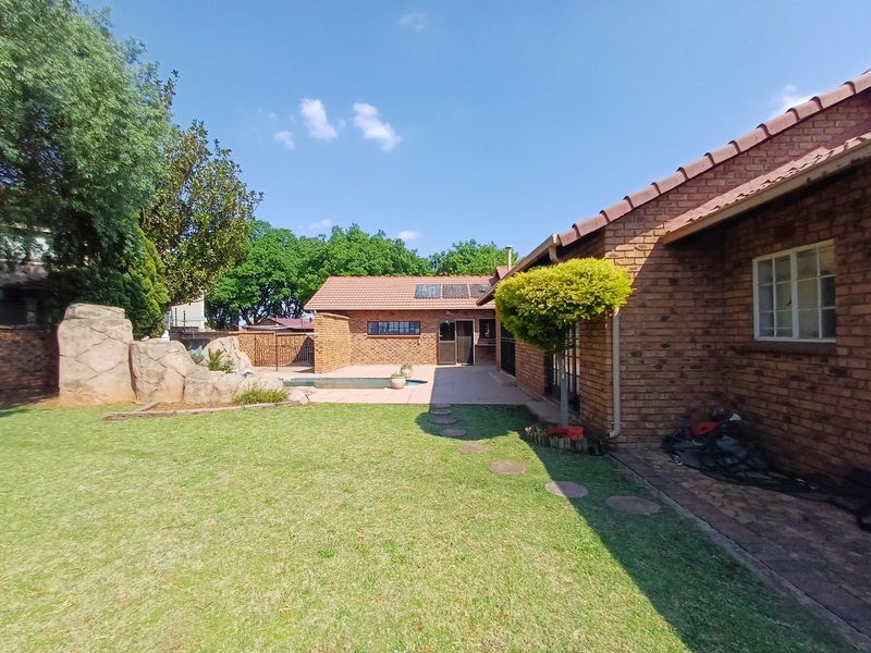 A spacious rear garden featuring a face brick house, a swimming pool, a paved patio area, and a decorative rock feature.