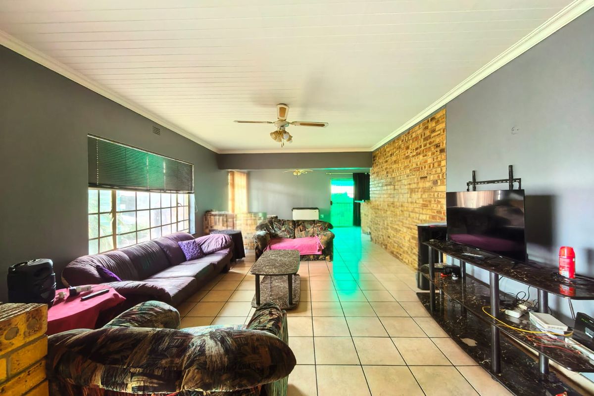 Spacious living room featuring a prominent exposed brick wall, tiled flooring, and a ceiling fan, offering a versatile layout for relaxation.