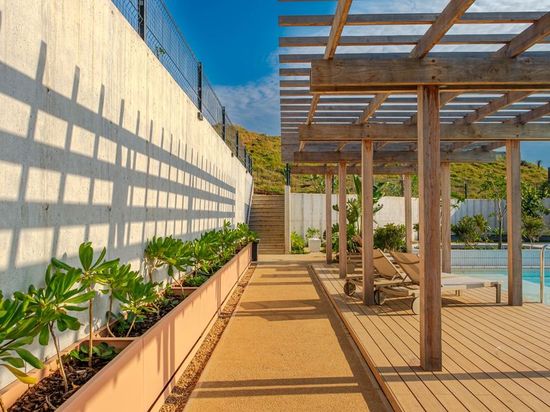 A well-maintained outdoor pool area featuring a timber pergola, wooden decking, concrete retaining walls, and integrated planter boxes.