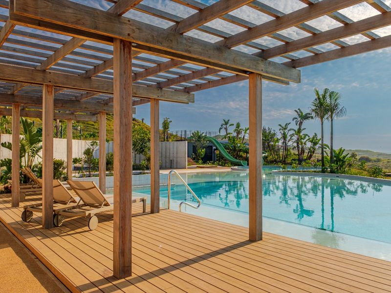 A well-maintained outdoor pool area featuring a timber pergola, wooden decking, and a swimming pool with a slide, set against a landscaped backdrop.
