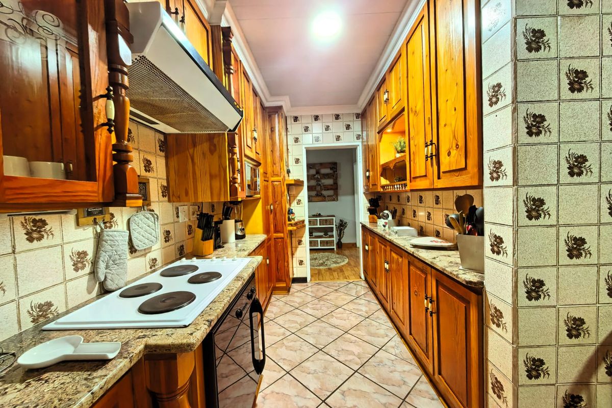 A galley-style kitchen featuring traditional wooden cabinetry, granite countertops, an electric hob, and a tiled backsplash with decorative floral accents.