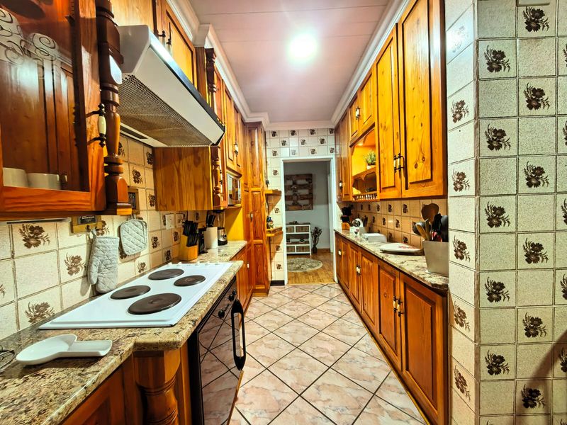 A galley-style kitchen featuring traditional wooden cabinetry, granite countertops, an electric hob, and a tiled backsplash with decorative floral accents.