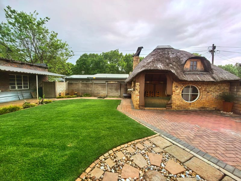 A well-maintained rear garden featuring a unique thatched-roof structure with a brick facade, paved patio area, and a neat lawn.