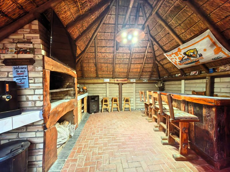 Rustic entertainment area featuring a thatched roof, exposed timber rafters, a built-in brick fireplace, and a custom wooden bar counter with brick flooring.