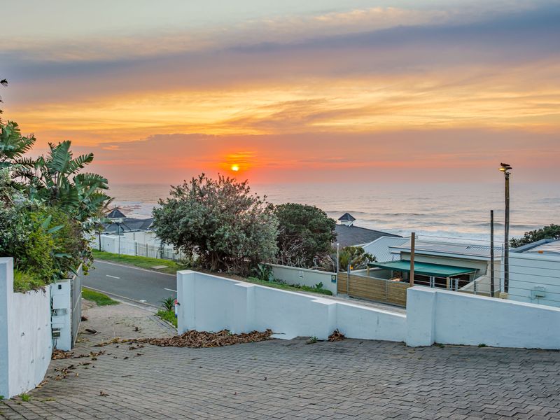 A scenic coastal view overlooking the ocean at sunset, featuring a paved driveway and boundary wall in the foreground.