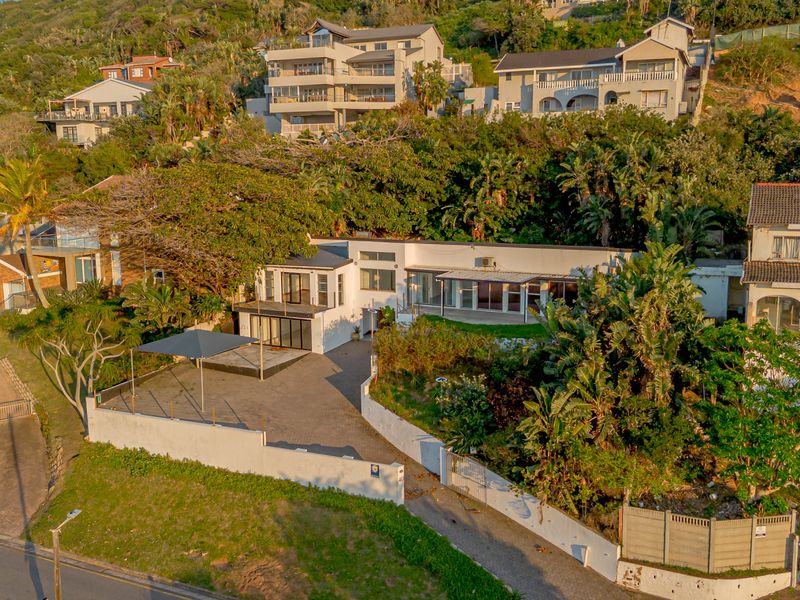 Aerial view of a residential property featuring a paved driveway, covered patio area, and established garden set against a hillside backdrop.