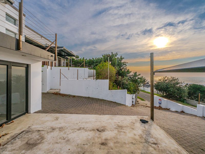 An outdoor rear area featuring a paved driveway, retaining walls, and ocean views, with a concrete patio space adjacent to the main building structure.
